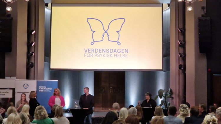 Stor skjerm på scene der det står "verdensdagen for psykisk helse". Under mange mennesker