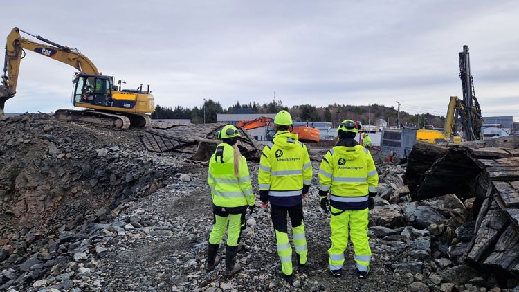 Tre personer står i gule arbeidsklær i et grustak med maskiner i nærheten