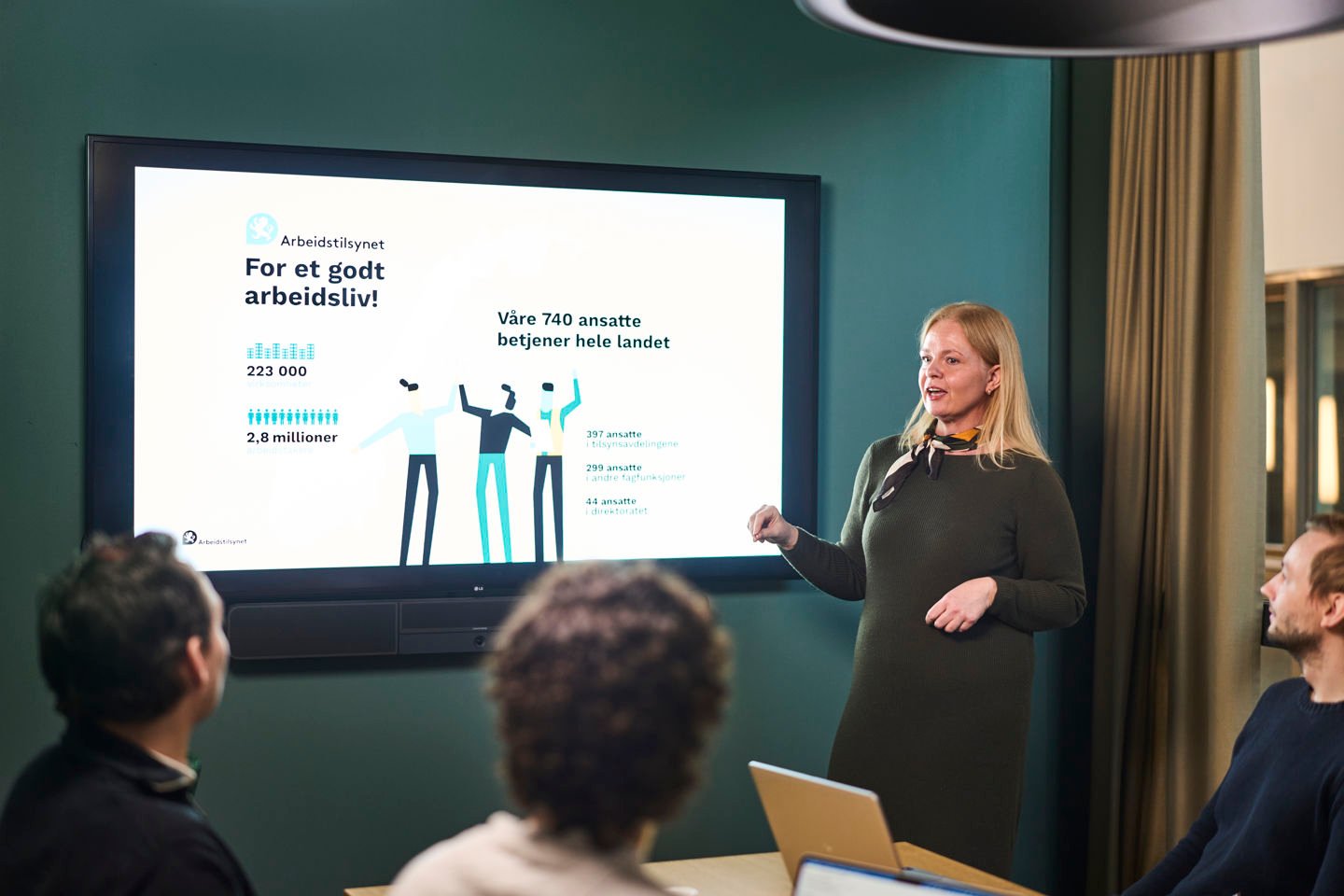 Ei dame står foran en skjerm med en presentasjon med fakta om Arbeidstilsynet. Vi ser tre tilhørere bakfra. 