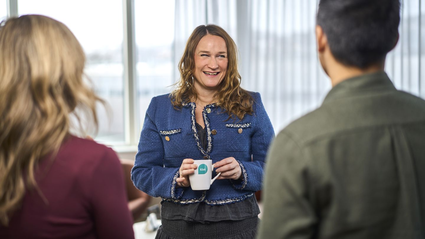 Dame med brunt langt hår står smilende med en kopp kaffe i hånda mens to mennsker vises bakfra